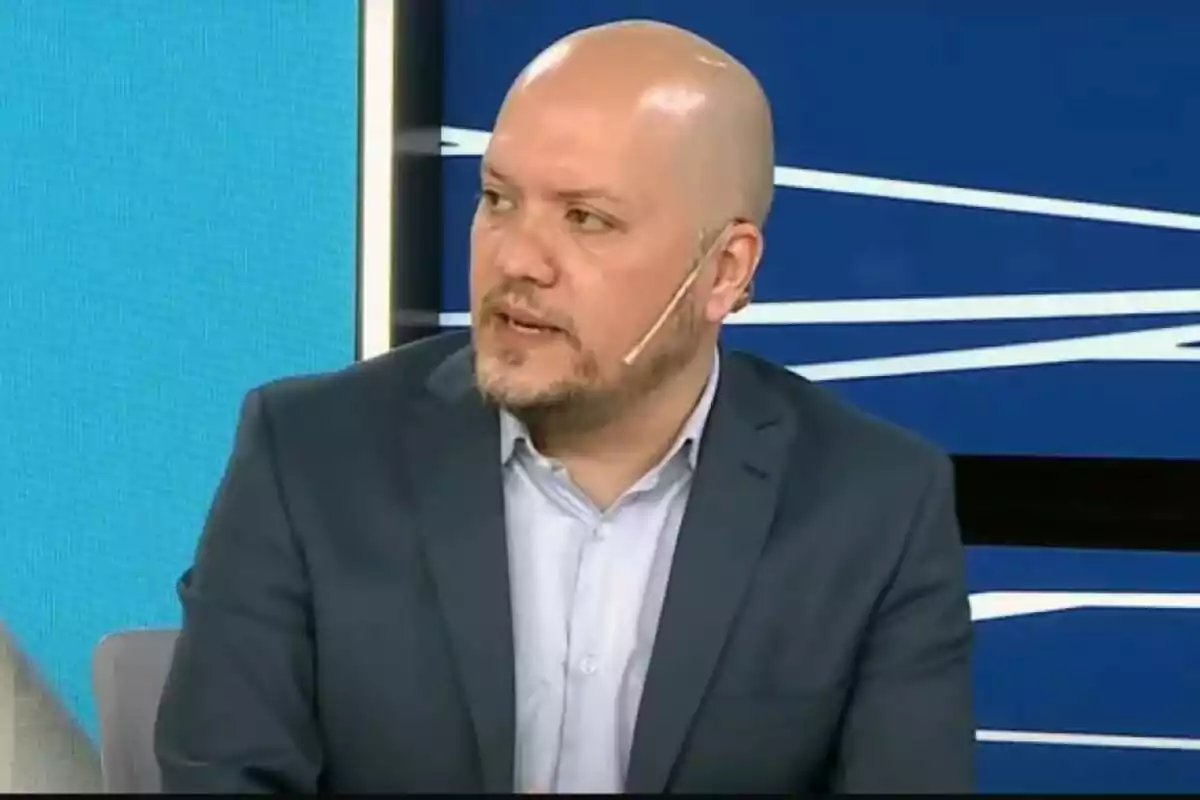 Hombre calvo con barba corta usando saco azul y camisa clara hablando en un set de televisión con fondo azul y micrófono en la mejilla Hombre calvo con barba corta usando saco azul y camisa clara hablando en un set de televisión con fondo azul y micrófono en la mejilla