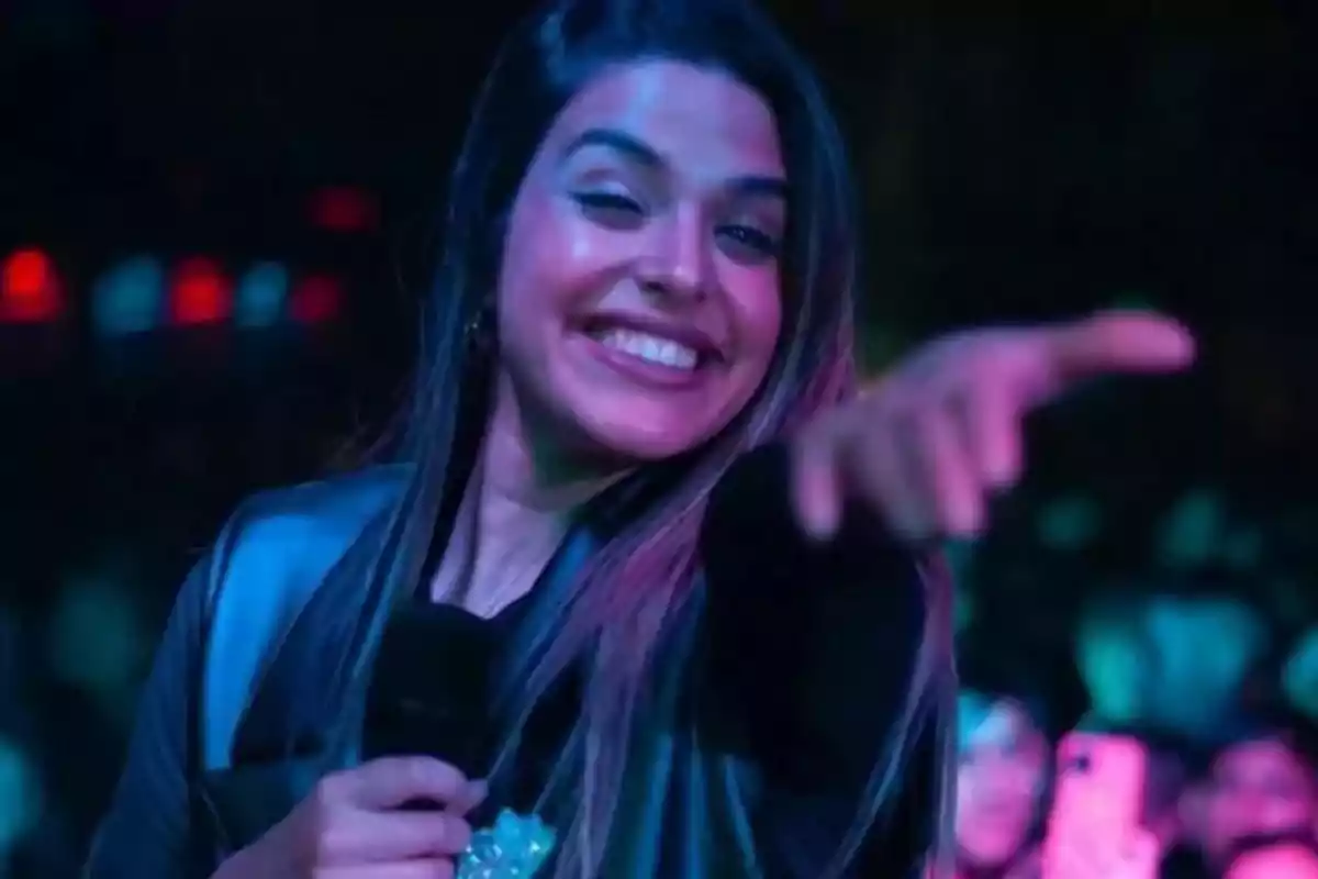 Smiling young woman with a microphone pointing at the camera in a concert setting with colorful lights Smiling young woman with a microphone pointing at the camera in a concert setting with colorful lights