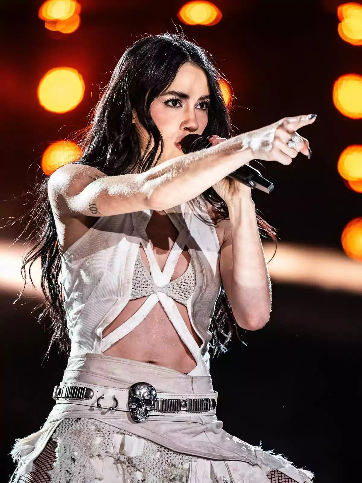 Long dark-haired singer dressed in white performing a song on stage while pointing toward the audience with blurred orange lights in the background Long dark-haired singer dressed in white performing a song on stage while pointing toward the audience with blurred orange lights in the background