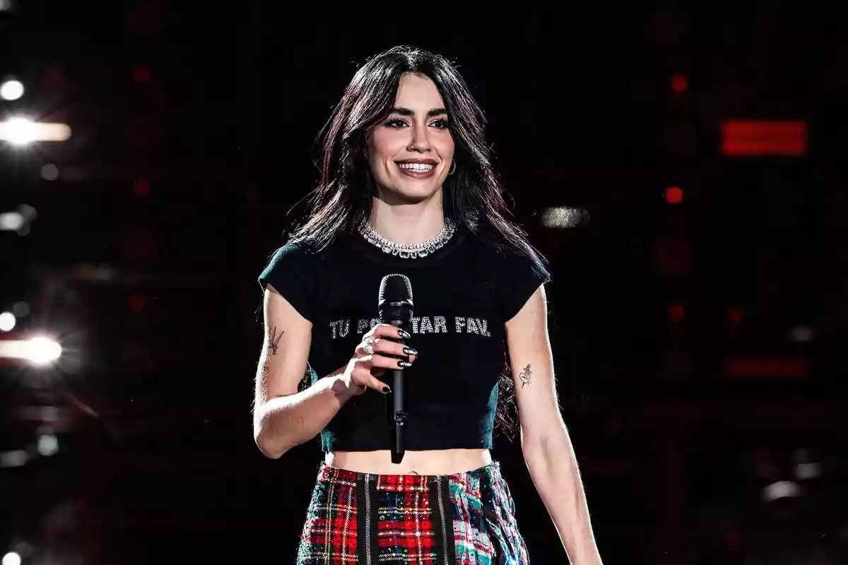 Young woman with long dark hair smiles while holding a microphone on stage, wearing a short black t-jersey and red plaid pants.