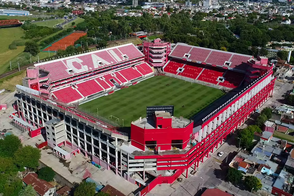 Vista aérea de un estadio de fútbol con gradas rojas y blancas rodeado de áreas verdes y edificios urbanos