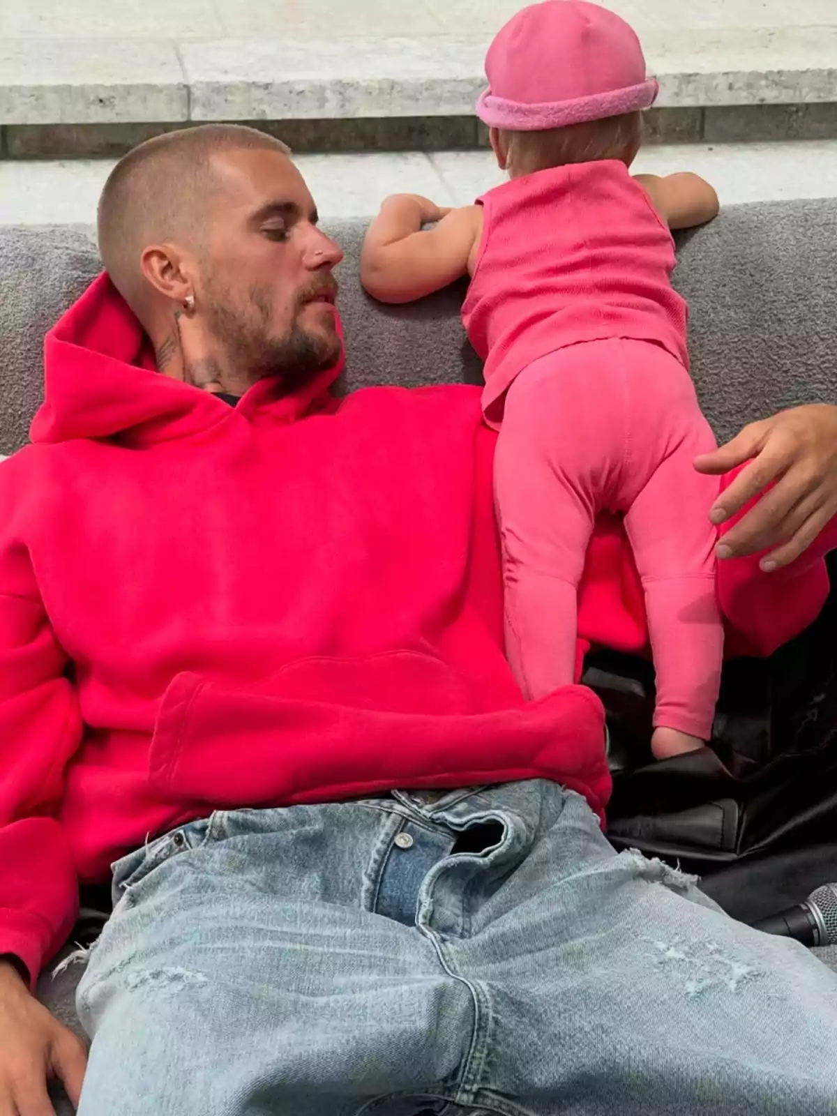 A man in a pink sweatjersey and ripped jeans is lying on a couch next to a baby dressed entirely in pink who is facing away. A man in a pink sweatjersey and ripped jeans is lying on a couch next to a baby dressed entirely in pink who is facing away.