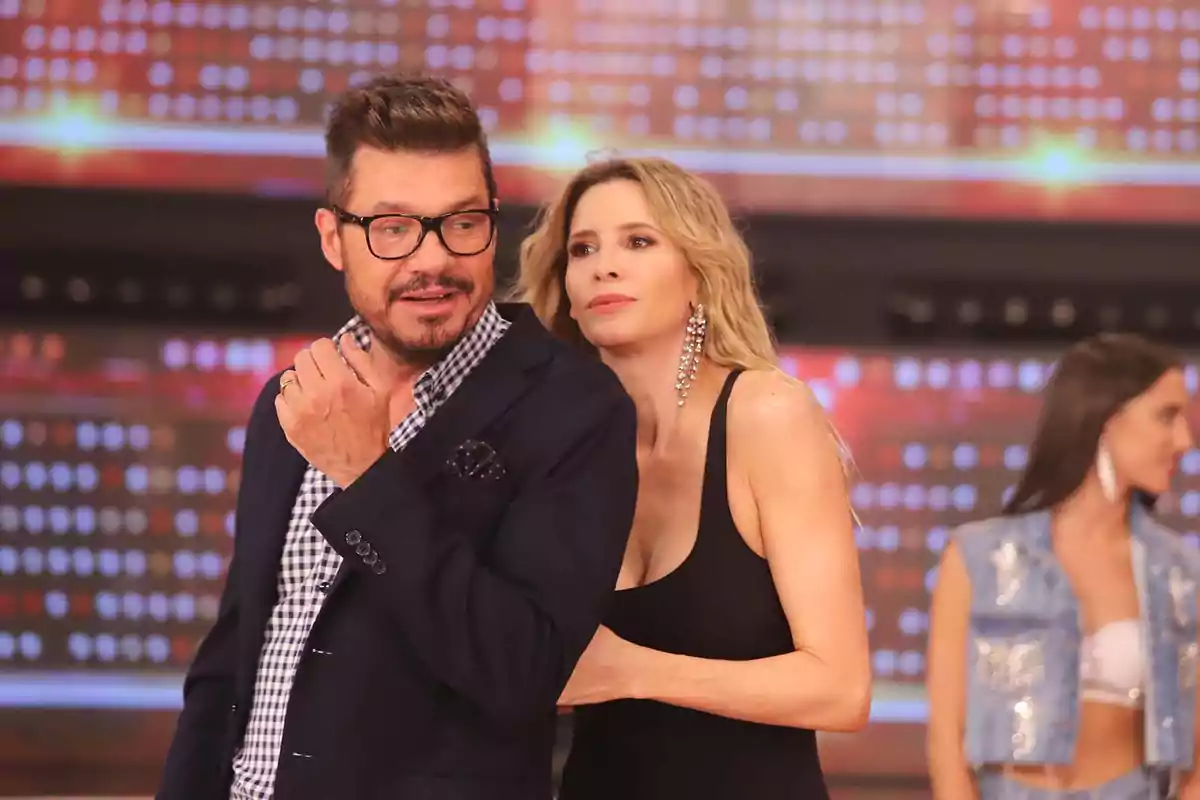 Un hombre con gafas y barba está de pie junto a una mujer rubia con vestido negro y grandes pendientes, ambos en un set de televisión con luces de fondo y otra mujer desenfocada al fondo. Un hombre con gafas y barba está de pie junto a una mujer rubia con vestido negro y grandes pendientes, ambos en un set de televisión con luces de fondo y otra mujer desenfocada al fondo.