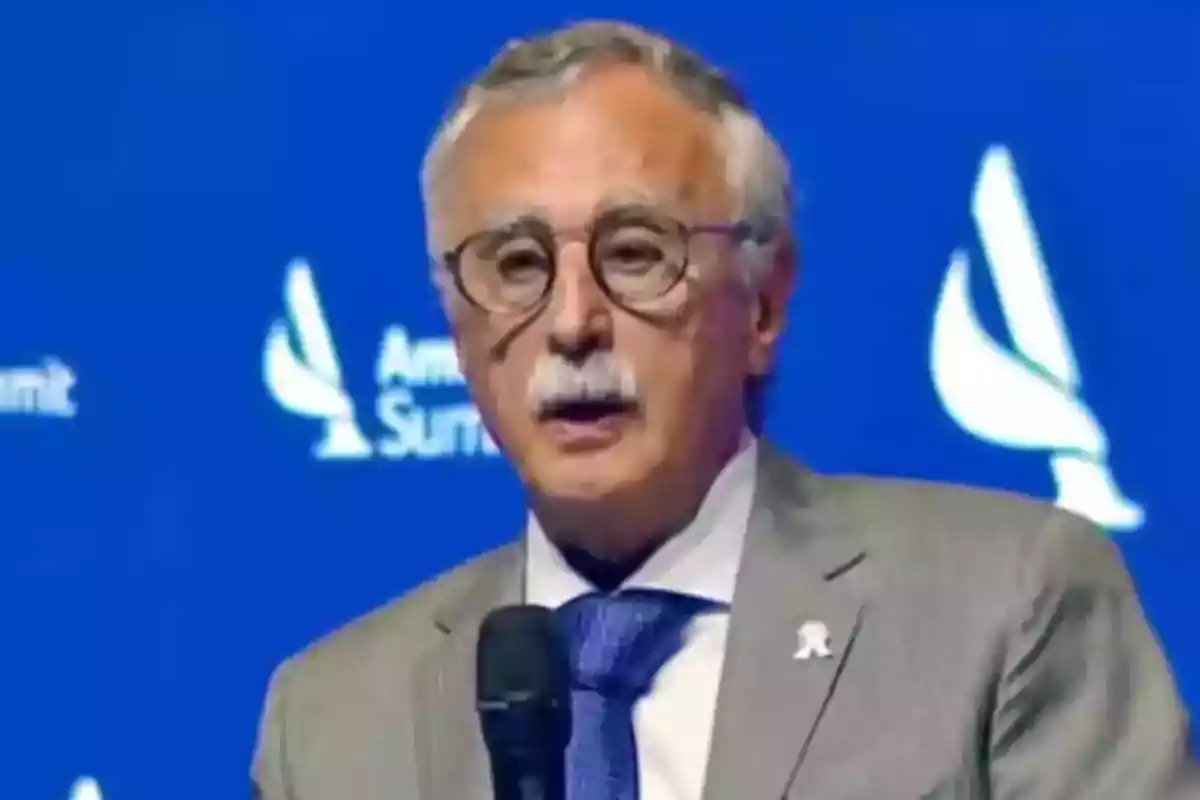 Older man with glasses and a mustache speaking at an event with a blue background Older man with glasses and a mustache speaking at an event with a blue background