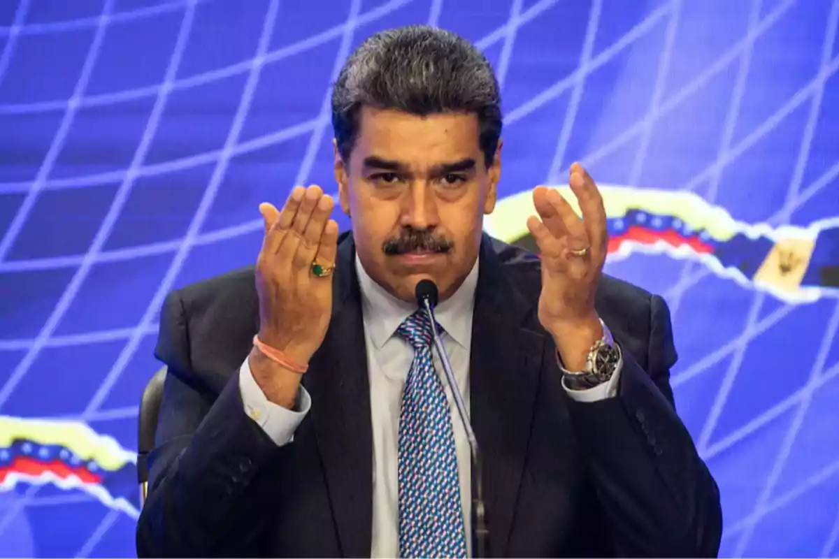Short-haired man with a mustache speaking in front of a microphone with his hands raised and a blue background with lines and a map. Short-haired man with a mustache speaking in front of a microphone with his hands raised and a blue background with lines and a map.