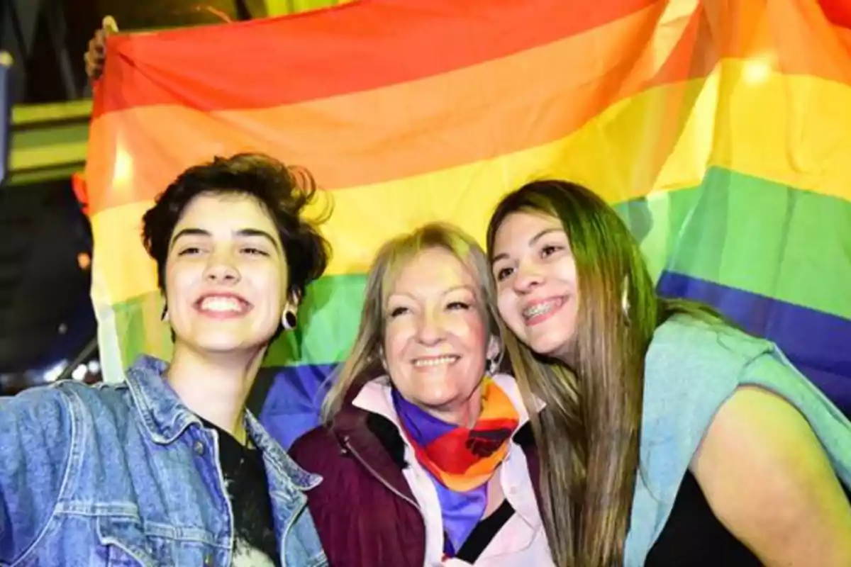 Tres personas sonríen mientras posan para una selfie frente a una bandera del orgullo LGBT Tres personas sonríen mientras posan para una selfie frente a una bandera del orgullo LGBT
