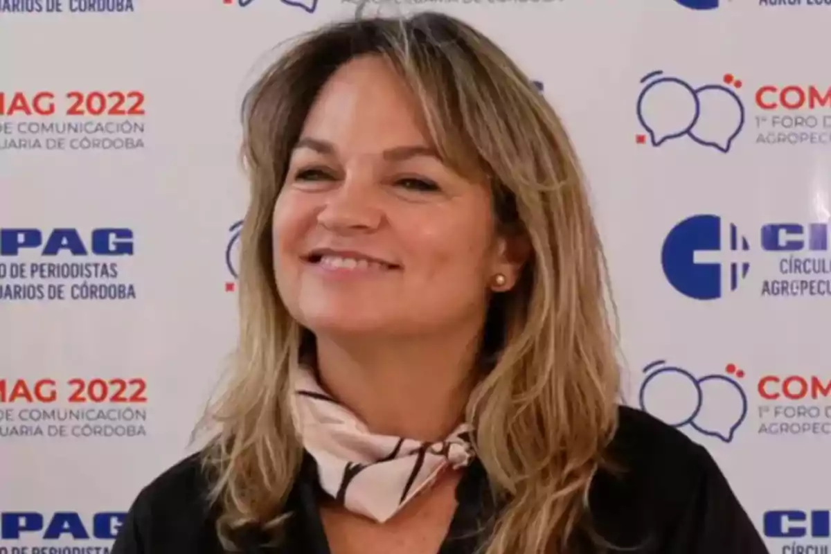 Smiling woman with blonde hair and a light scarf around her neck posing in front of a backdrop with logos from an agricultural communication event Smiling woman with blonde hair and a light scarf around her neck posing in front of a backdrop with logos from an agricultural communication event