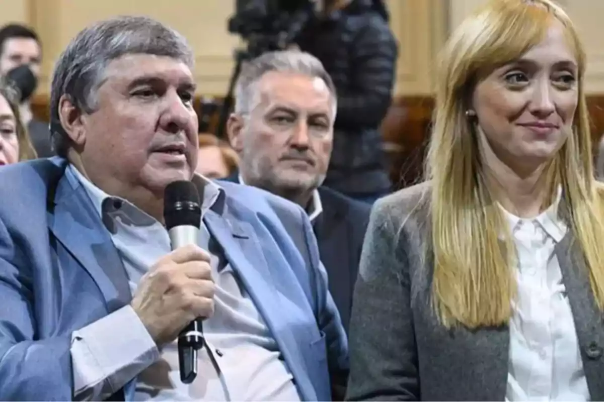 A gray-haired man holds a microphone while sitting next to a blonde woman who is smiling slightly at a formal event with people in the background. A gray-haired man holds a microphone while sitting next to a blonde woman who is smiling slightly at a formal event with people in the background.