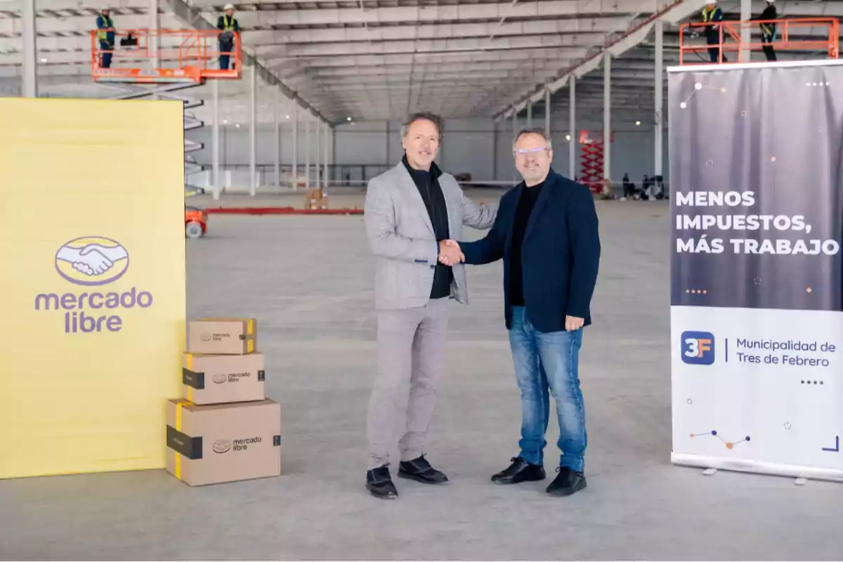 Dos hombres se dan la mano en el centro de un gran galpón en construcción, junto a cajas de Mercado Libre y un cartel de la Municipalidad de Tres de Febrero que dice menos impuestos más trabajo Dos hombres se dan la mano en el centro de un gran galpón en construcción, junto a cajas de Mercado Libre y un cartel de la Municipalidad de Tres de Febrero que dice menos impuestos más trabajo