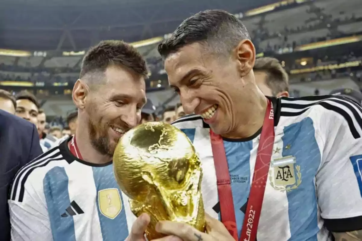 Two Argentine players smile and celebrate while holding the World Cup trophy in a crowded stadium. Two Argentine players smile and celebrate while holding the World Cup trophy in a crowded stadium.