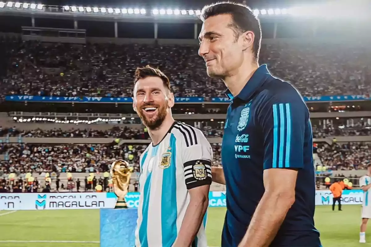 Dos personas sonríen en un estadio de fútbol lleno de público, una de ellas lleva la camiseta de la selección argentina y la otra viste ropa deportiva azul, al fondo se observa el trofeo de la Copa del Mundo.