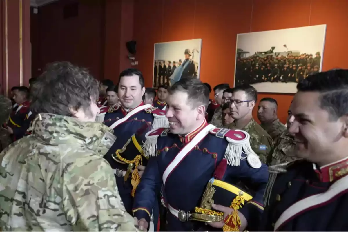 A group of military personnel in dress uniforms smile and greet a person in a camouflage jacket in a room decorated with historical photographs on the wall. A group of military personnel in dress uniforms smile and greet a person in a camouflage jacket in a room decorated with historical photographs on the wall.