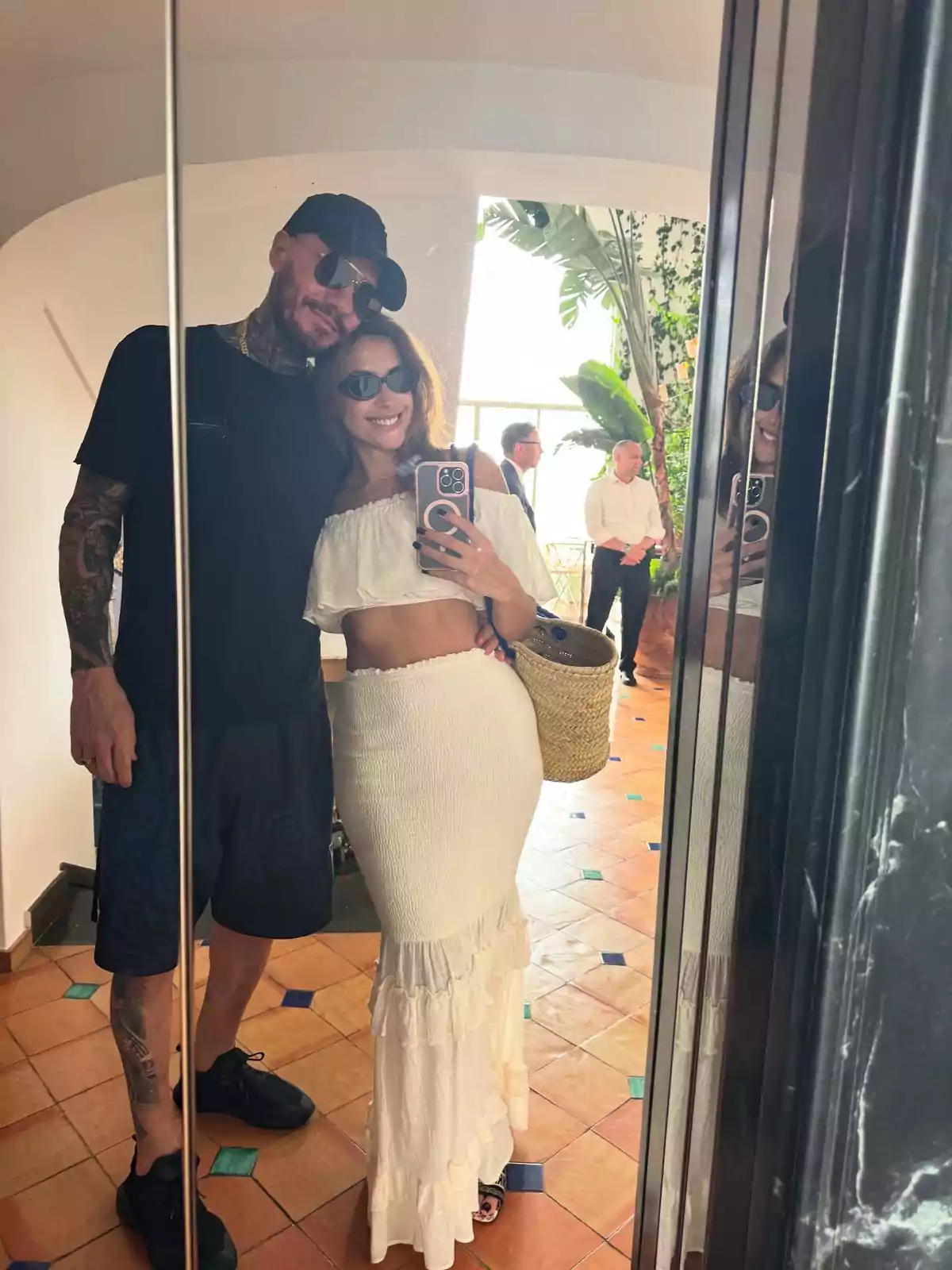 Couple taking a selfie in front of a mirror in a well-lit place with plants and people in the background, both wearing sunglasses and summer clothes Couple taking a selfie in front of a mirror in a well-lit place with plants and people in the background, both wearing sunglasses and summer clothes