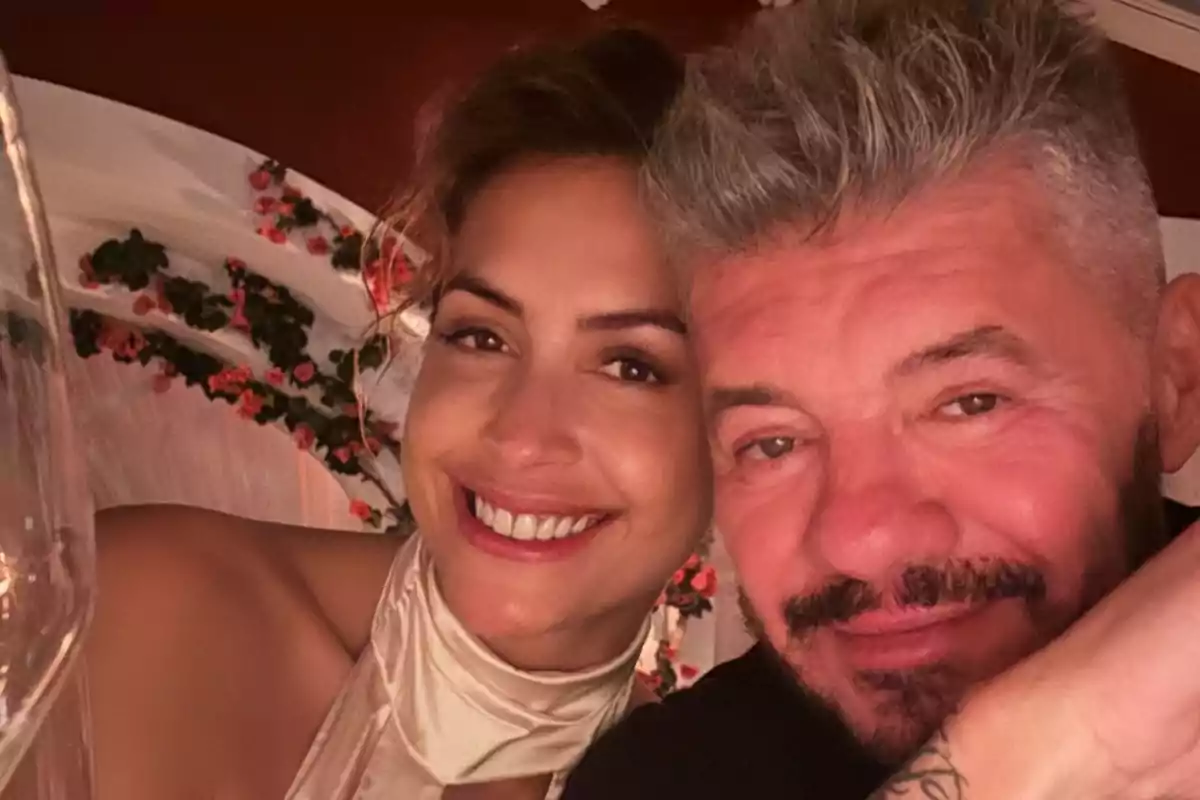 A couple smiles, hugging each other while posing for a selfie in a flower-decorated setting.