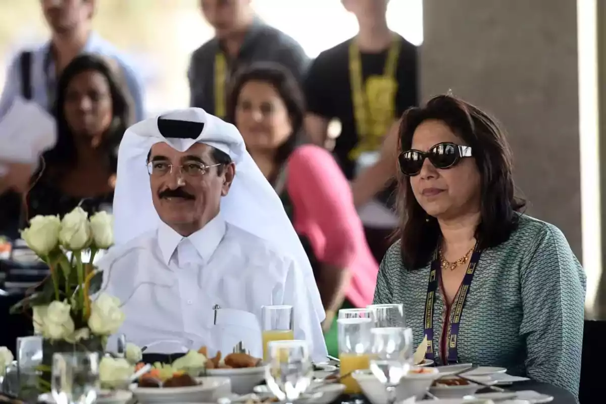 Dos personas sentadas en una mesa elegante con comida y flores blancas, rodeadas de otras personas al fondo Dos personas sentadas en una mesa elegante con comida y flores blancas, rodeadas de otras personas al fondo