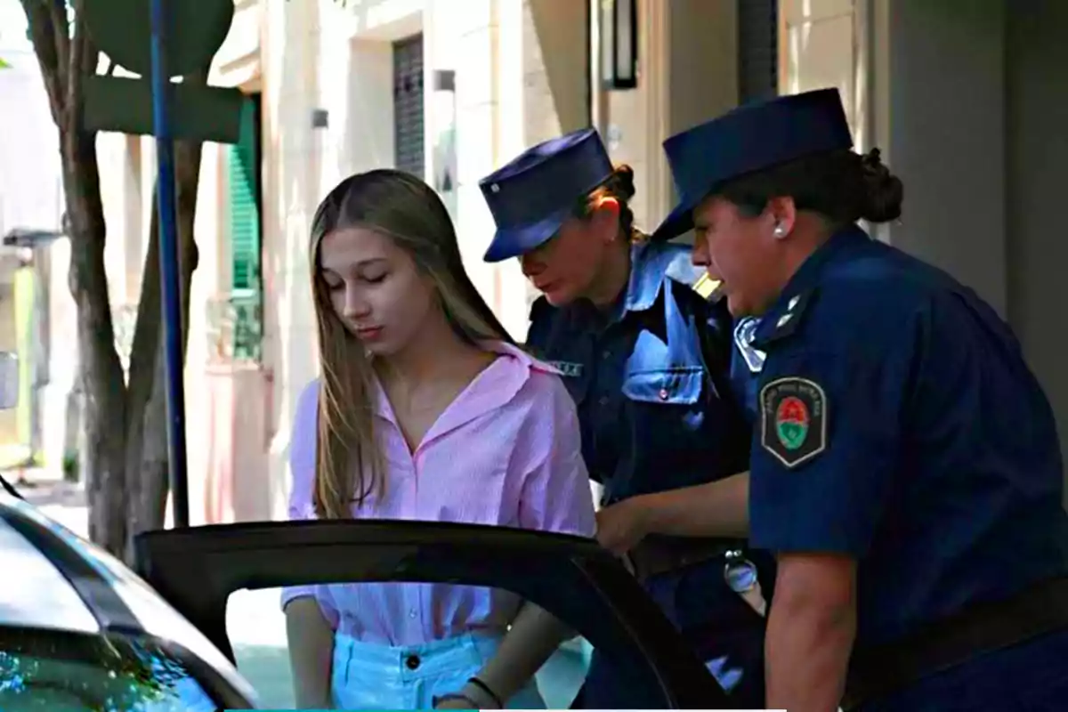 A young woman in a pink jersey is escorted by two police officers toward a patrol car on a residential street during the day. A young woman in a pink jersey is escorted by two police officers toward a patrol car on a residential street during the day.