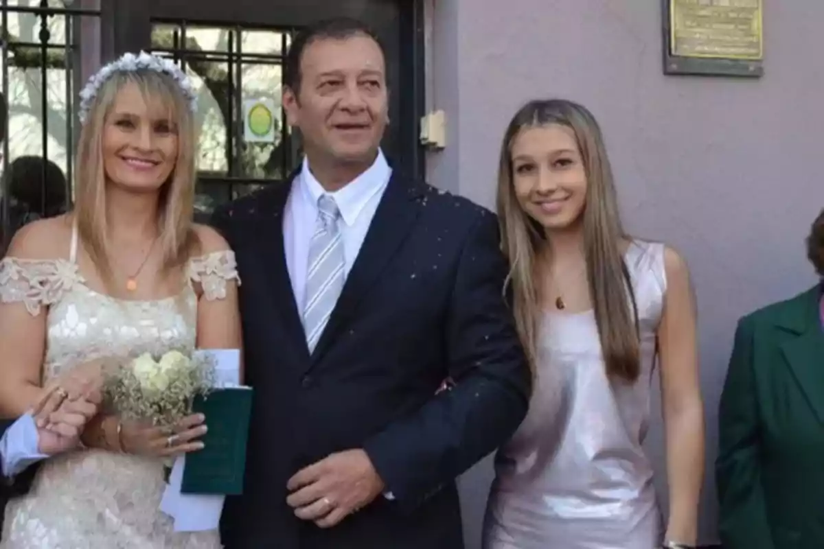 Three people smile and pose together at what appears to be a wedding; the woman on the left is wearing a white dress and holding a bouquet, the man in the center is dressed in a dark suit, and the young woman on the right is wearing a light-colored dress. Three people smile and pose together at what appears to be a wedding; the woman on the left is wearing a white dress and holding a bouquet, the man in the center is dressed in a dark suit, and the young woman on the right is wearing a light-colored dress.