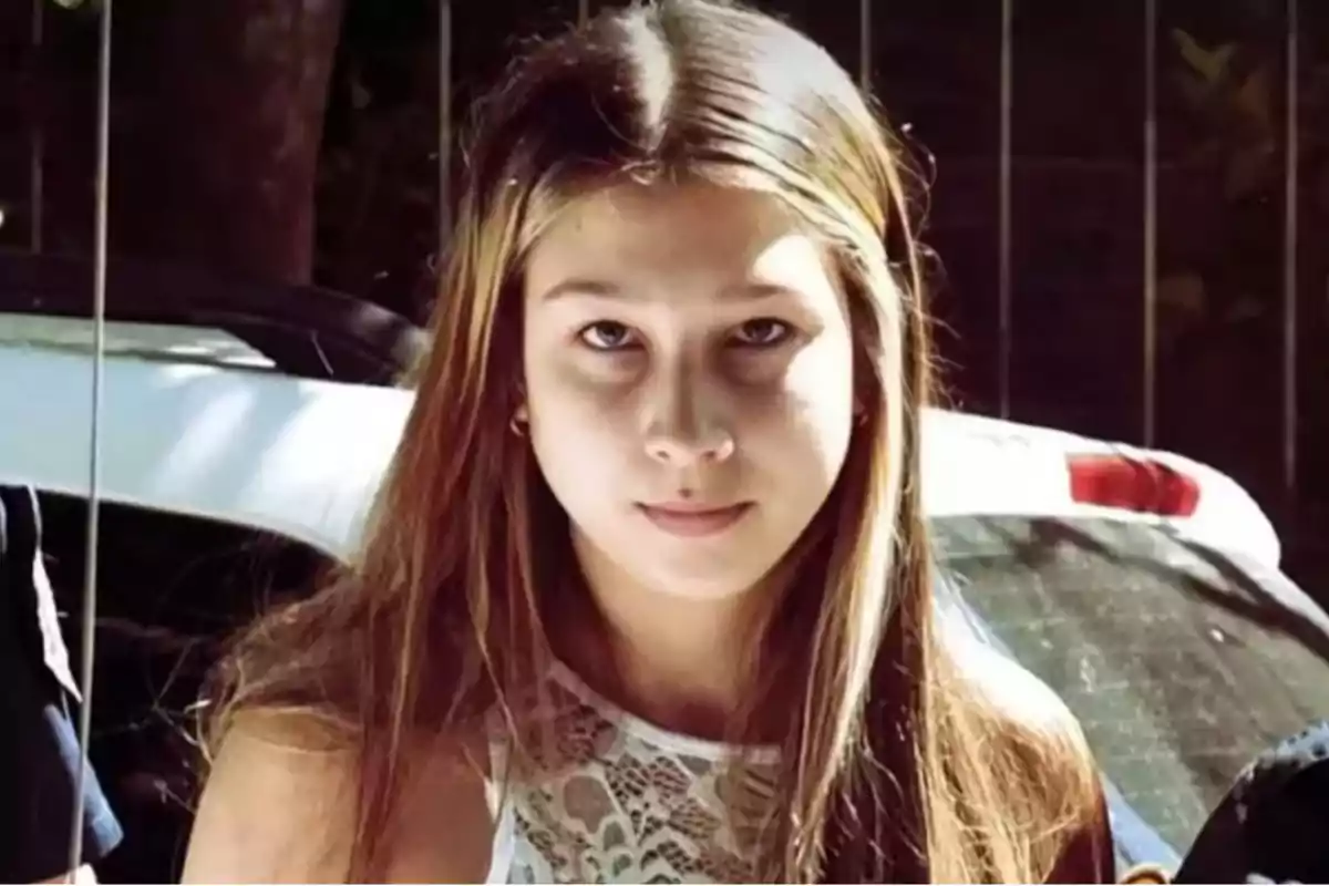 Young person with long, straight hair and a serious expression looking at the camera in an outdoor setting with cars in the background