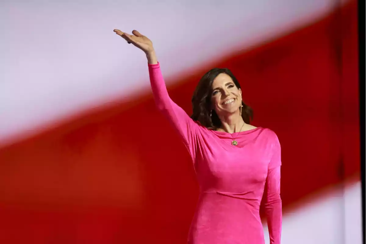 Mujer con vestido rosa saludando y sonriendo frente a un fondo rojo y blanco difuminado