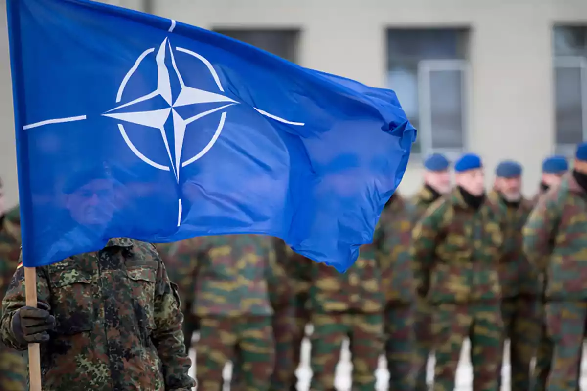 Soldado sosteniendo una bandera de la OTAN frente a un grupo de militares con uniforme de camuflaje y boinas azules Soldado sosteniendo una bandera de la OTAN frente a un grupo de militares con uniforme de camuflaje y boinas azules