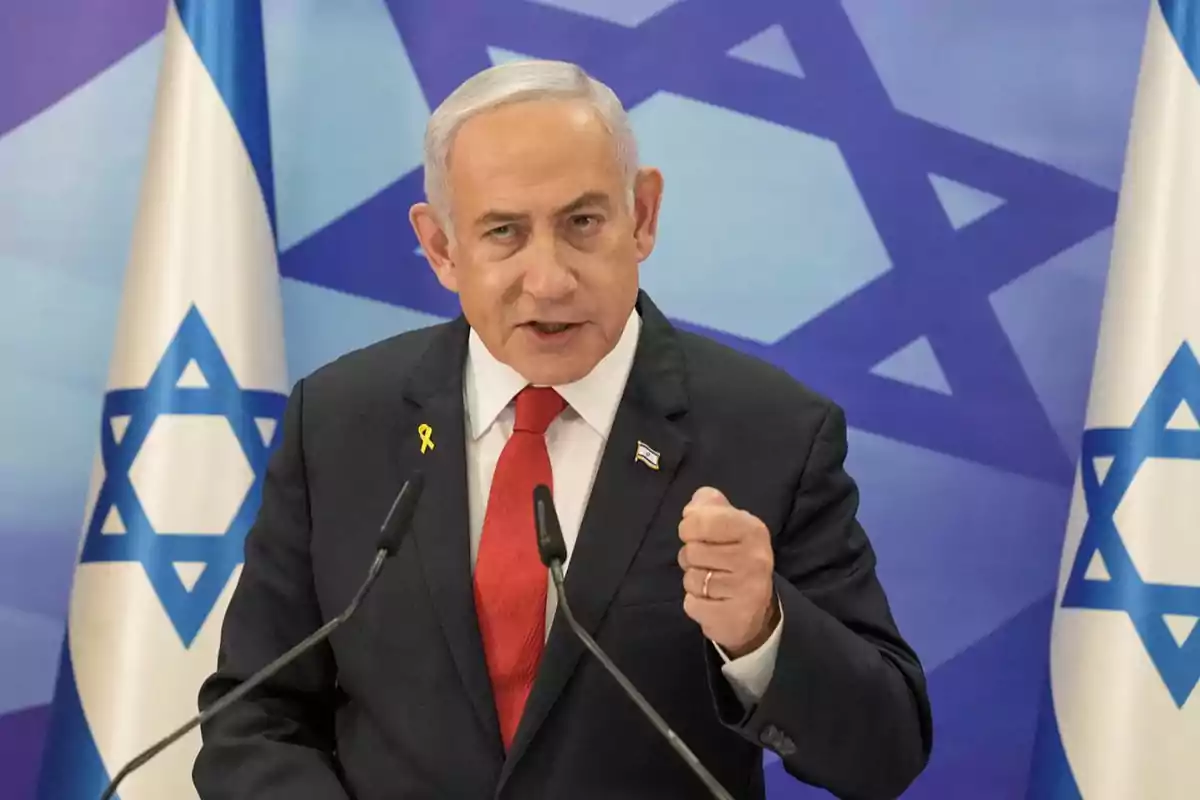 Gray-haired man in a dark suit and red tie speaking in front of microphones with Israeli flags in the background Gray-haired man in a dark suit and red tie speaking in front of microphones with Israeli flags in the background