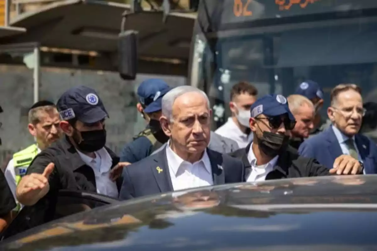 A gray-haired man in a dark suit is escorted by security agents wearing black caps and masks as he walks with a group of people in front of a bus.