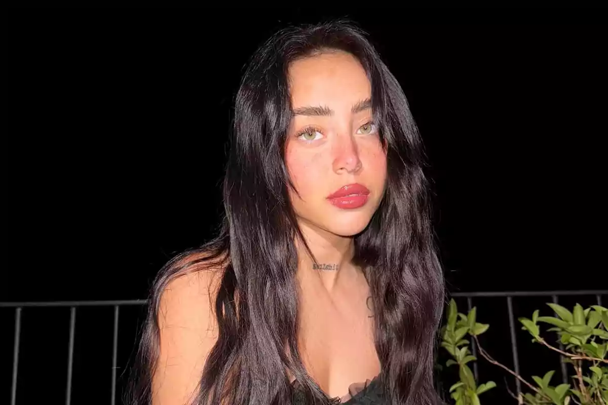 Long-haired, dark-haired woman with red lips and freckles, posing outdoors at night in front of a black background and some plants.