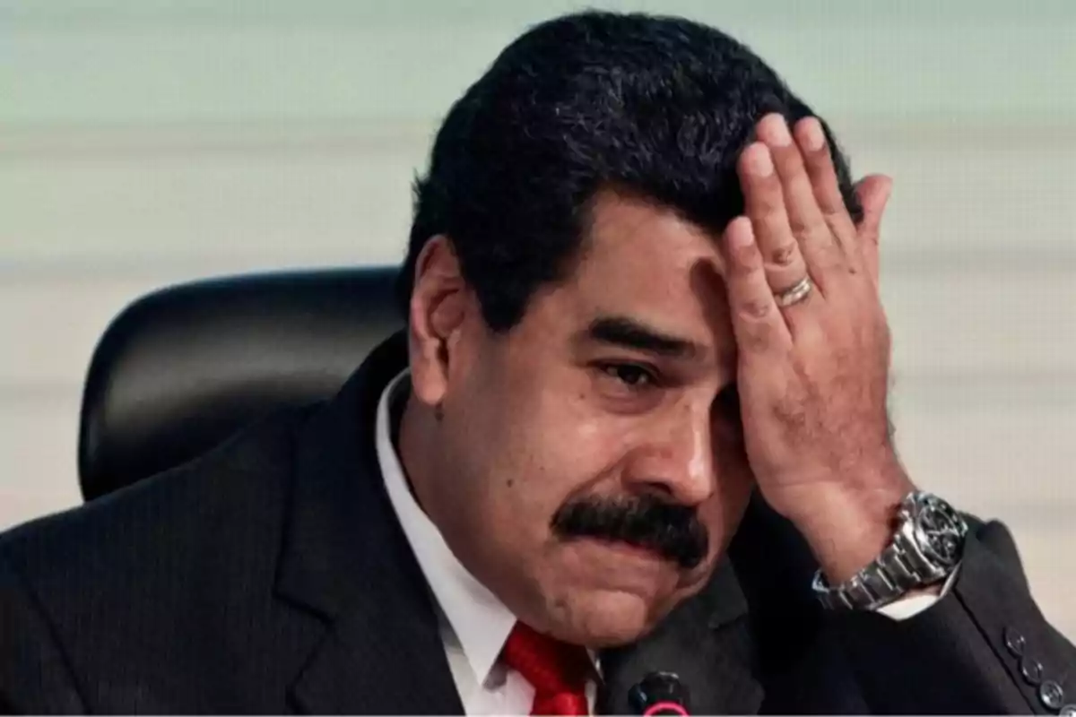 Man with a mustache and dark suit sitting, with a worried expression and his hand on his forehead