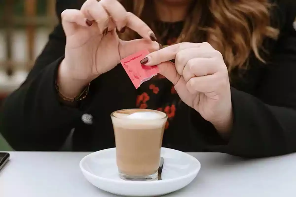 Person pouring a packet of sweetener into a cup of coffee with milk Person pouring a packet of sweetener into a cup of coffee with milk