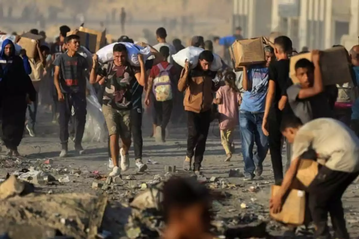 A group of people walks across a dusty, debris-filled area while carrying boxes and bags on their shoulders. A group of people walks across a dusty, debris-filled area while carrying boxes and bags on their shoulders.