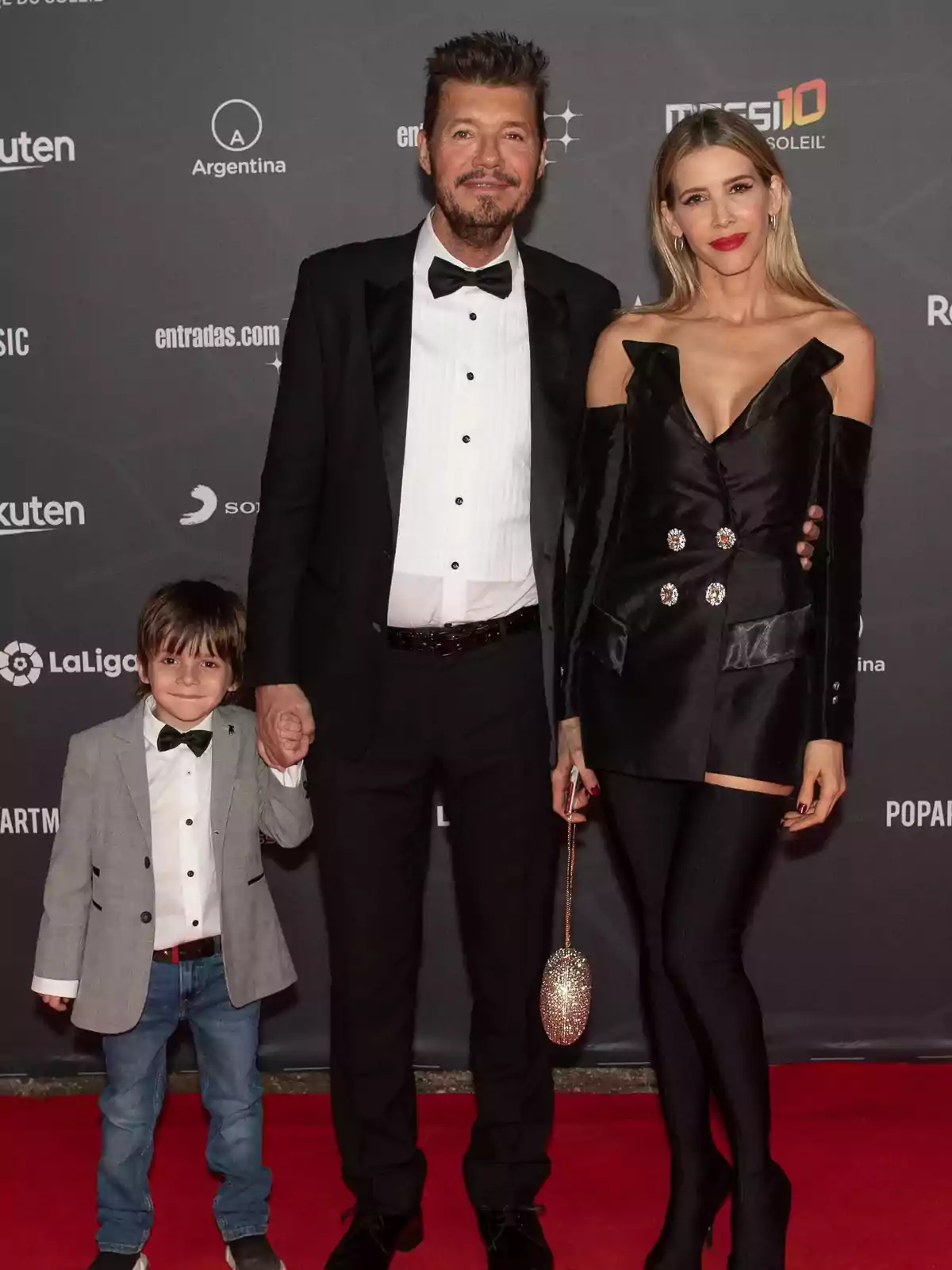 Una familia posa en una alfombra roja con fondo de logotipos, el hombre lleva esmoquin, la mujer viste un conjunto negro elegante y el niño lleva saco gris y moño Una familia posa en una alfombra roja con fondo de logotipos, el hombre lleva esmoquin, la mujer viste un conjunto negro elegante y el niño lleva saco gris y moño