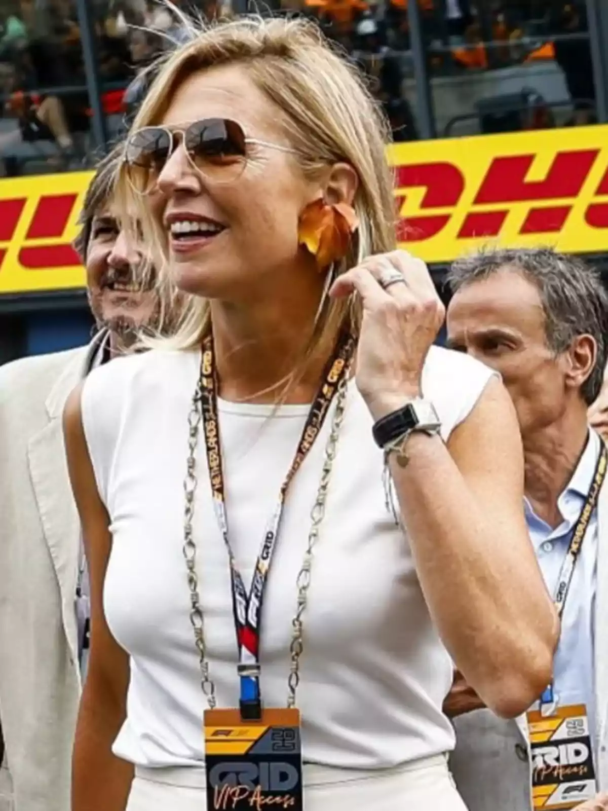 Blonde woman with sunglasses and large earrings smiles while attending a motorsport event with a VIP pass hanging around her neck Blonde woman with sunglasses and large earrings smiles while attending a motorsport event with a VIP pass hanging around her neck