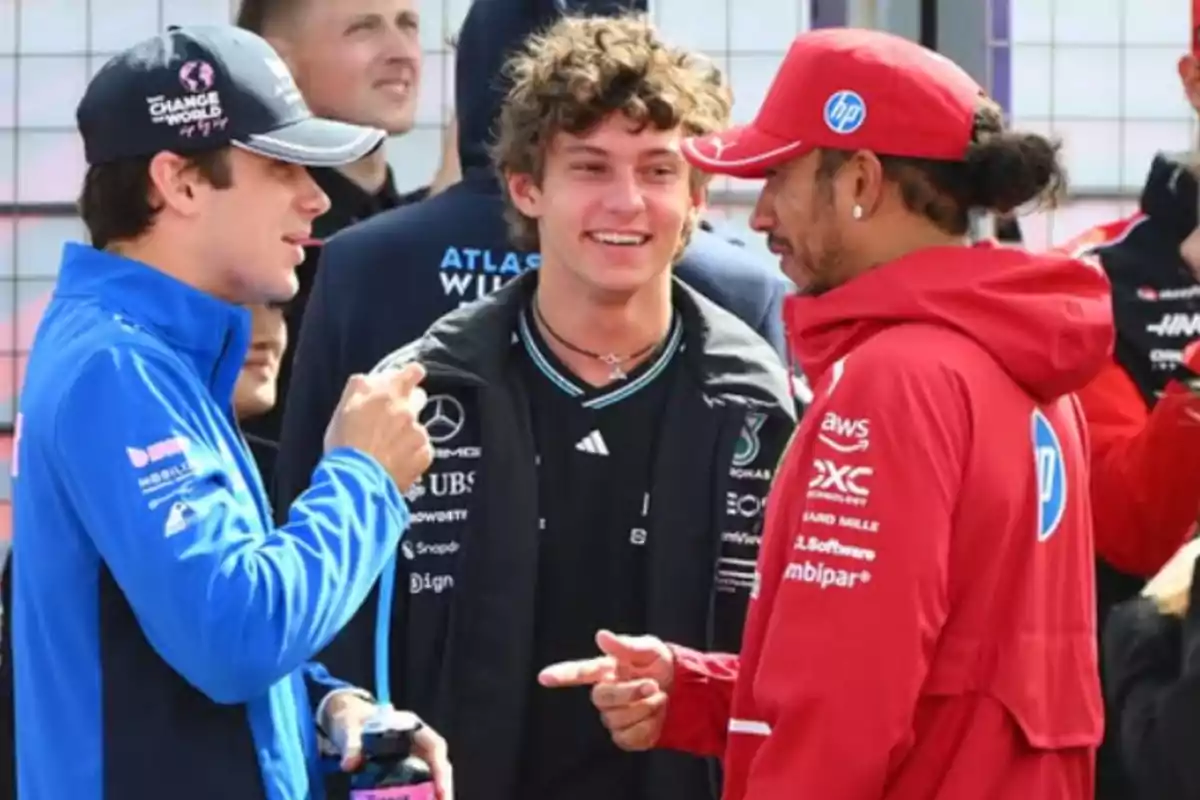 Three people are talking and smiling in a sports setting; one is wearing a blue jacket, another a black jacket, and the third a red jacket with a red cap. Three people are talking and smiling in a sports setting; one is wearing a blue jacket, another a black jacket, and the third a red jacket with a red cap.