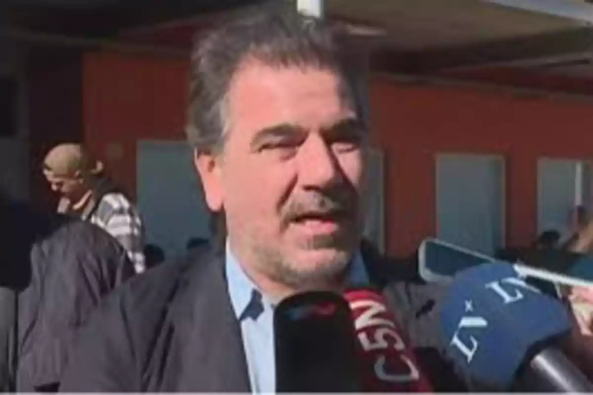 Gray-haired man speaking in front of several press microphones in an outdoor space