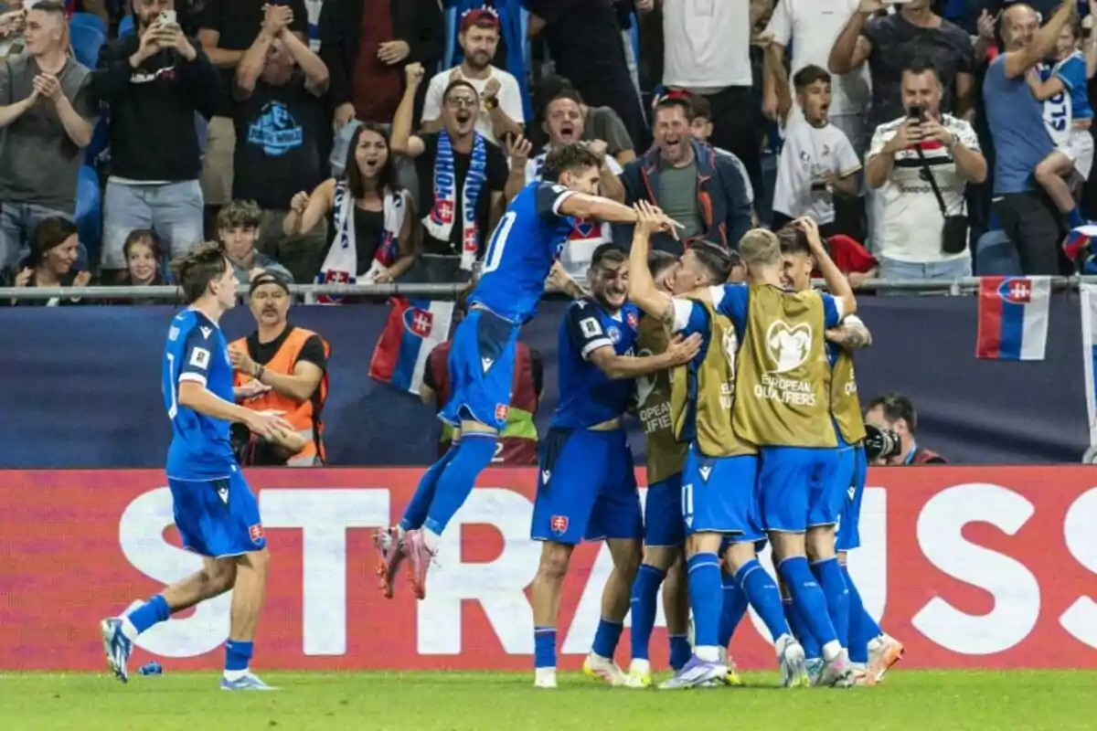 Jugadores de fútbol celebran un gol abrazándose frente a una multitud de aficionados que aplauden y animan en las gradas, con banderas de Eslovaquia visibles en el fondo. Jugadores de fútbol celebran un gol abrazándose frente a una multitud de aficionados que aplauden y animan en las gradas, con banderas de Eslovaquia visibles en el fondo.