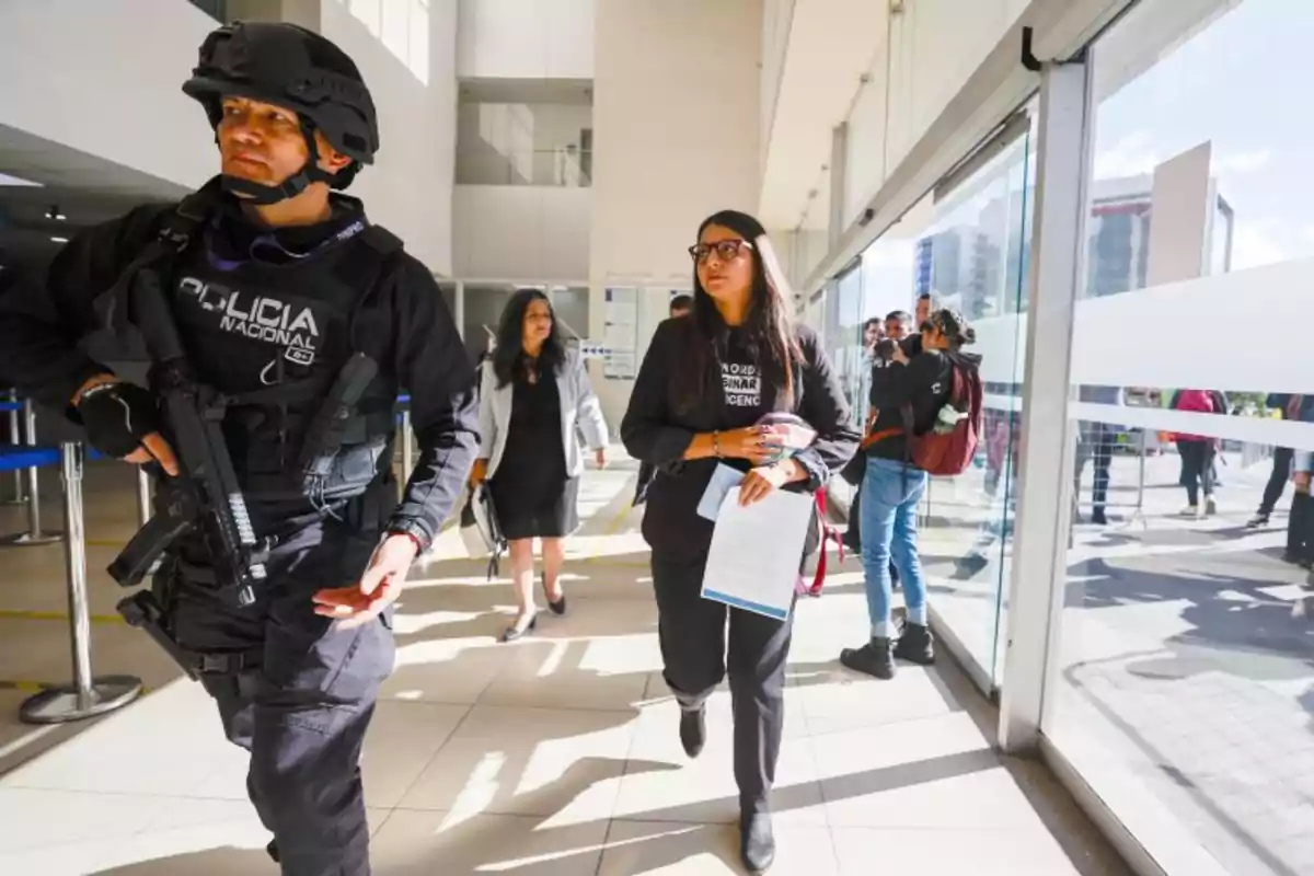 A heavily armed police officer walks alongside several people inside a modern building with glass walls and natural light. A heavily armed police officer walks alongside several people inside a modern building with glass walls and natural light.