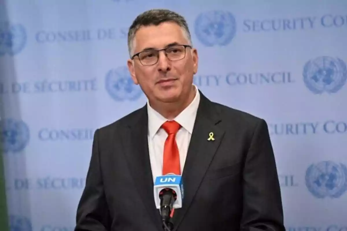 Man with glasses and a dark suit speaking in front of a UN microphone with the Security Council in the background Man with glasses and a dark suit speaking in front of a UN microphone with the Security Council in the background