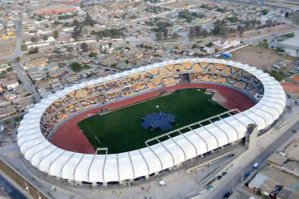 Vista aérea de un estadio de fútbol con techo blanco y graderías amarillas rodeado de una zona urbana Vista aérea de un estadio de fútbol con techo blanco y graderías amarillas rodeado de una zona urbana