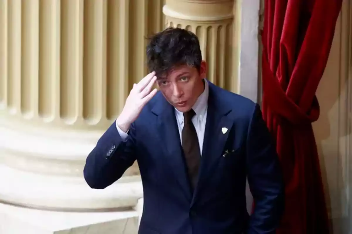 Short dark-haired person in a blue suit and dark tie raising their hand to their head in an elegant setting with columns and red curtains Short dark-haired person in a blue suit and dark tie raising their hand to their head in an elegant setting with columns and red curtains