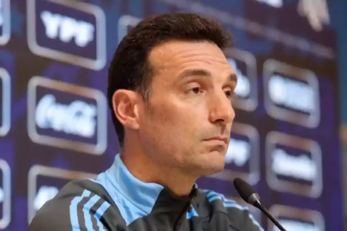 Hombre con chaqueta deportiva celeste y negra hablando en una conferencia de prensa con fondo de logotipos desenfocados Hombre con chaqueta deportiva celeste y negra hablando en una conferencia de prensa con fondo de logotipos desenfocados