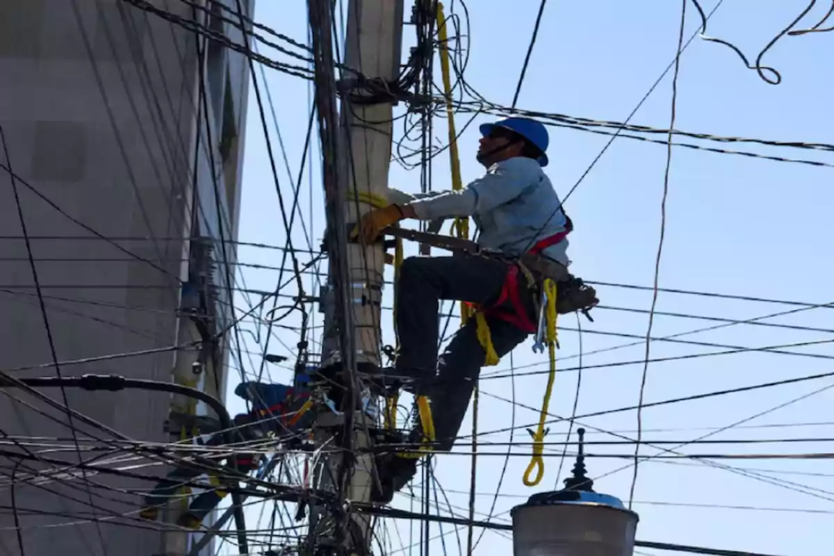 Worker with a blue helmet and harness climbing a pole surrounded by electrical wires performing maintenance work Worker with a blue helmet and harness climbing a pole surrounded by electrical wires performing maintenance work