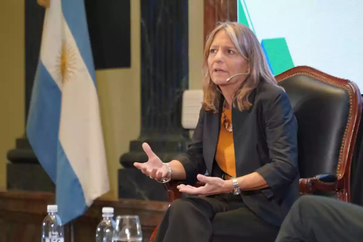Woman sitting and speaking with a headset microphone at a formal event with the Argentine flag in the background Woman sitting and speaking with a headset microphone at a formal event with the Argentine flag in the background