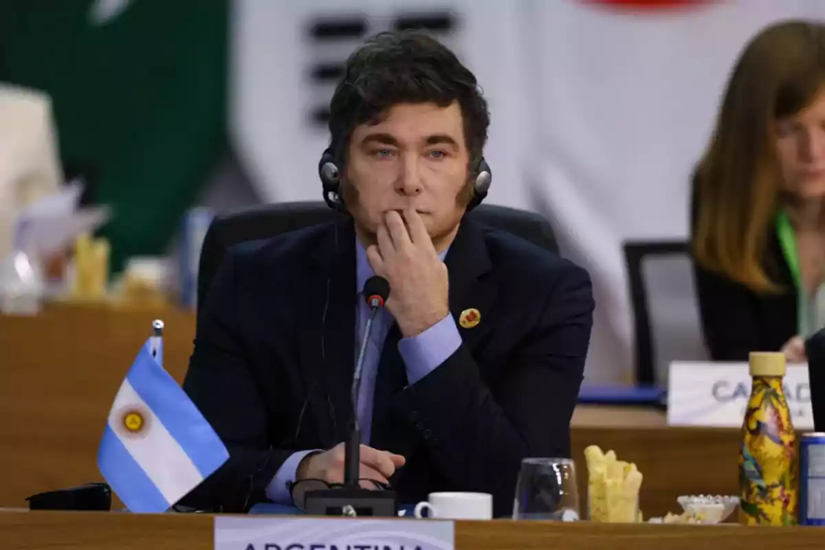 A man wearing headphones and a dark suit is sitting at a conference table next to an Argentine flag, with a thoughtful expression and his hand on his face. A man wearing headphones and a dark suit is sitting at a conference table next to an Argentine flag, with a thoughtful expression and his hand on his face.