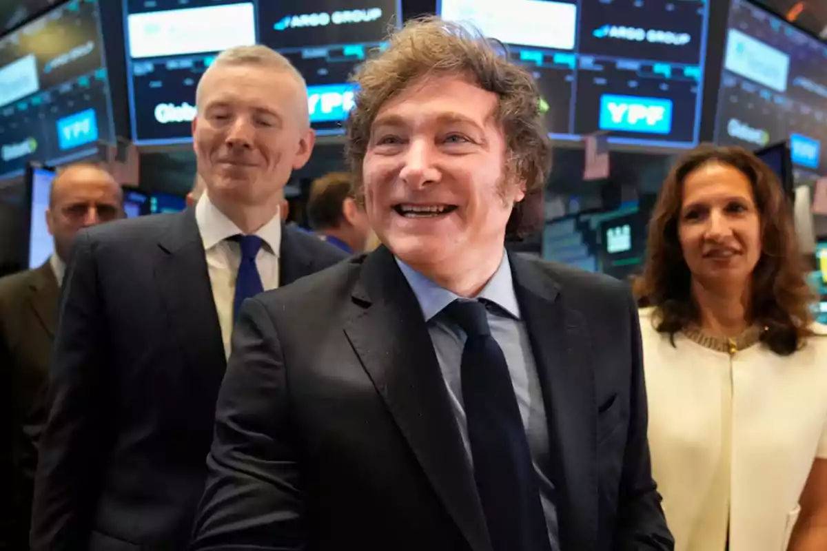 Un hombre sonriente con traje oscuro y corbata azul está rodeado de otras personas en un ambiente de bolsa de valores con pantallas electrónicas de fondo. Un hombre sonriente con traje oscuro y corbata azul está rodeado de otras personas en un ambiente de bolsa de valores con pantallas electrónicas de fondo.
