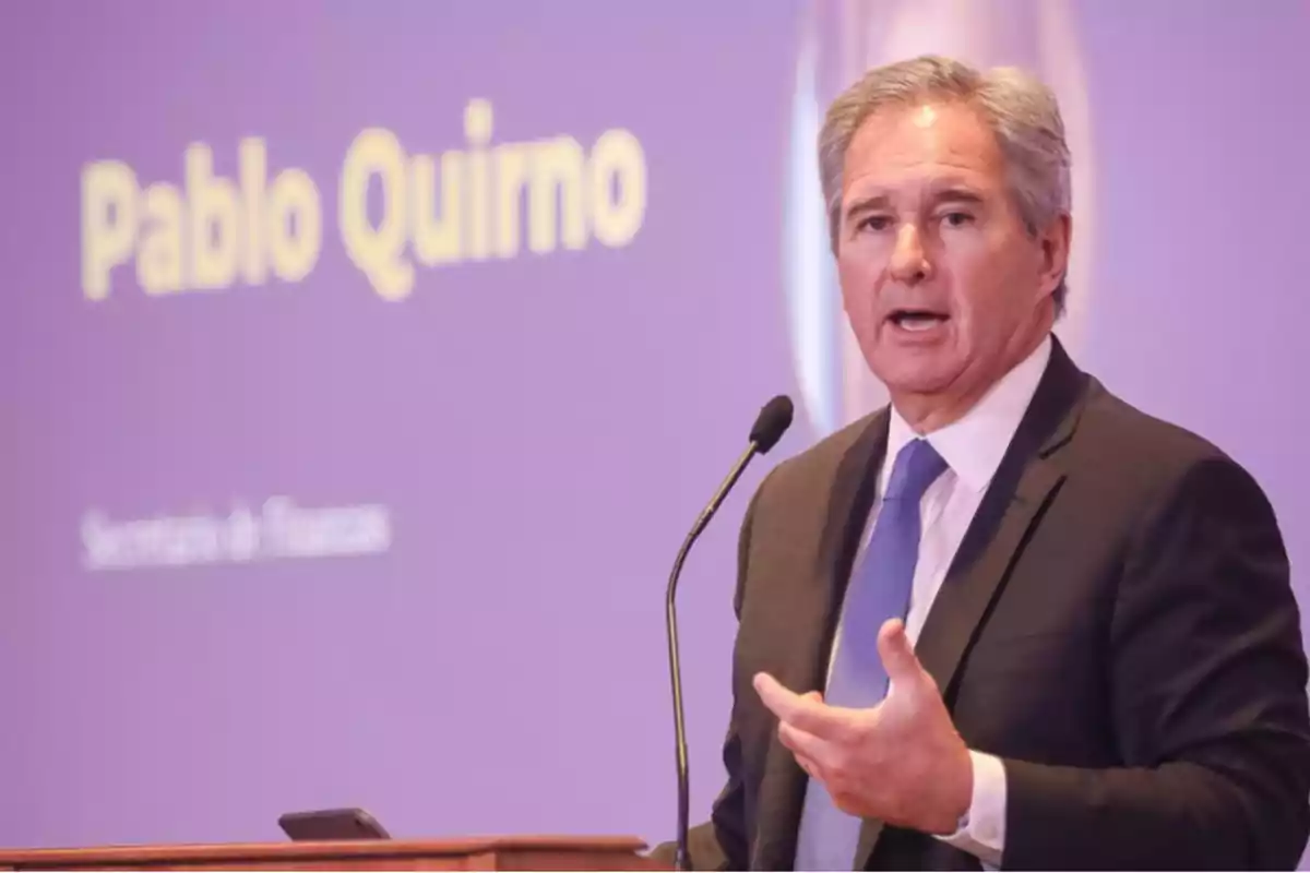 Hombre de cabello canoso con traje y corbata azul hablando frente a un micrófono con una pantalla de fondo que muestra el nombre Pablo Quirno Hombre de cabello canoso con traje y corbata azul hablando frente a un micrófono con una pantalla de fondo que muestra el nombre Pablo Quirno