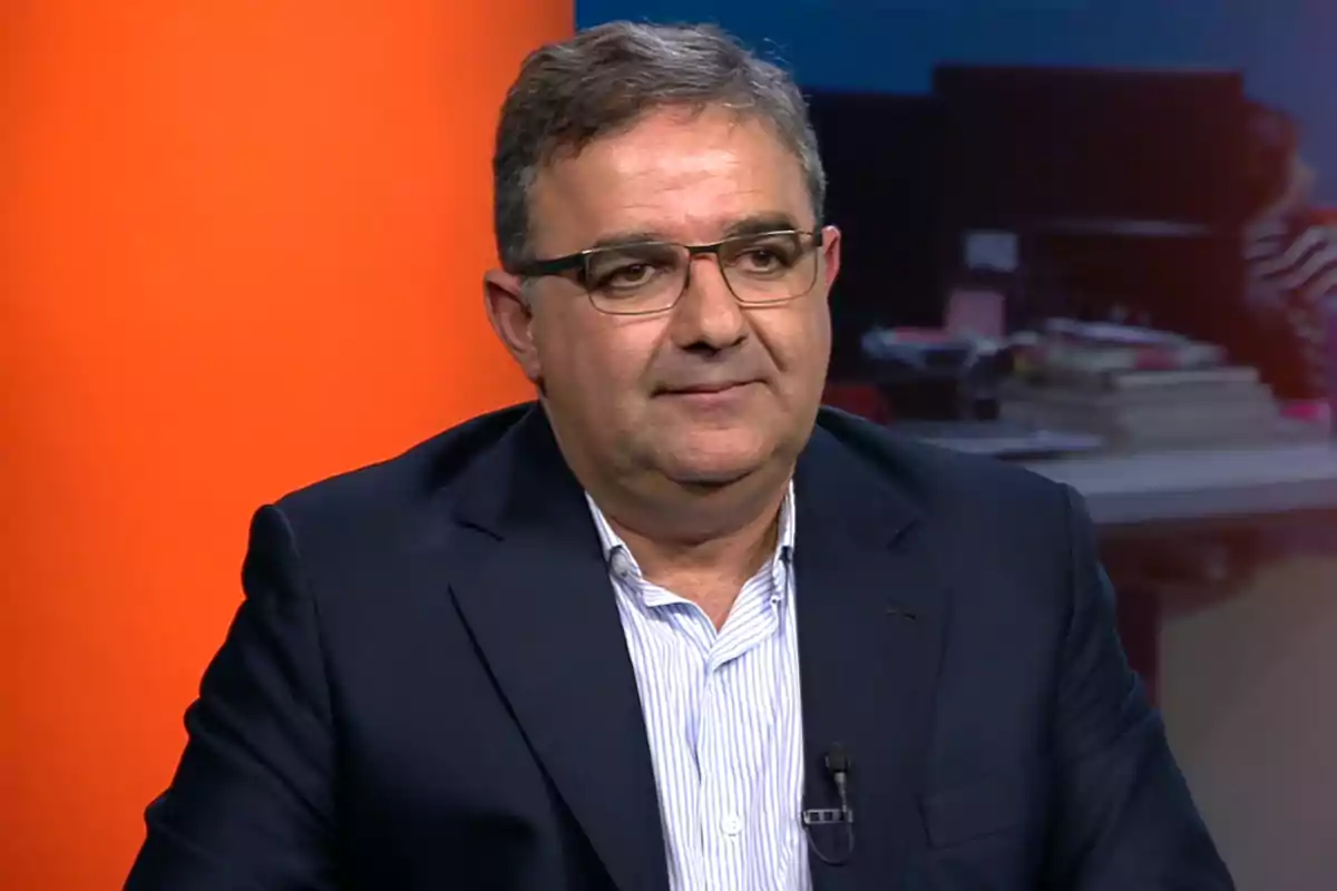 Middle-aged man with glasses and a dark jacket sitting in a studio with an orange and blue background