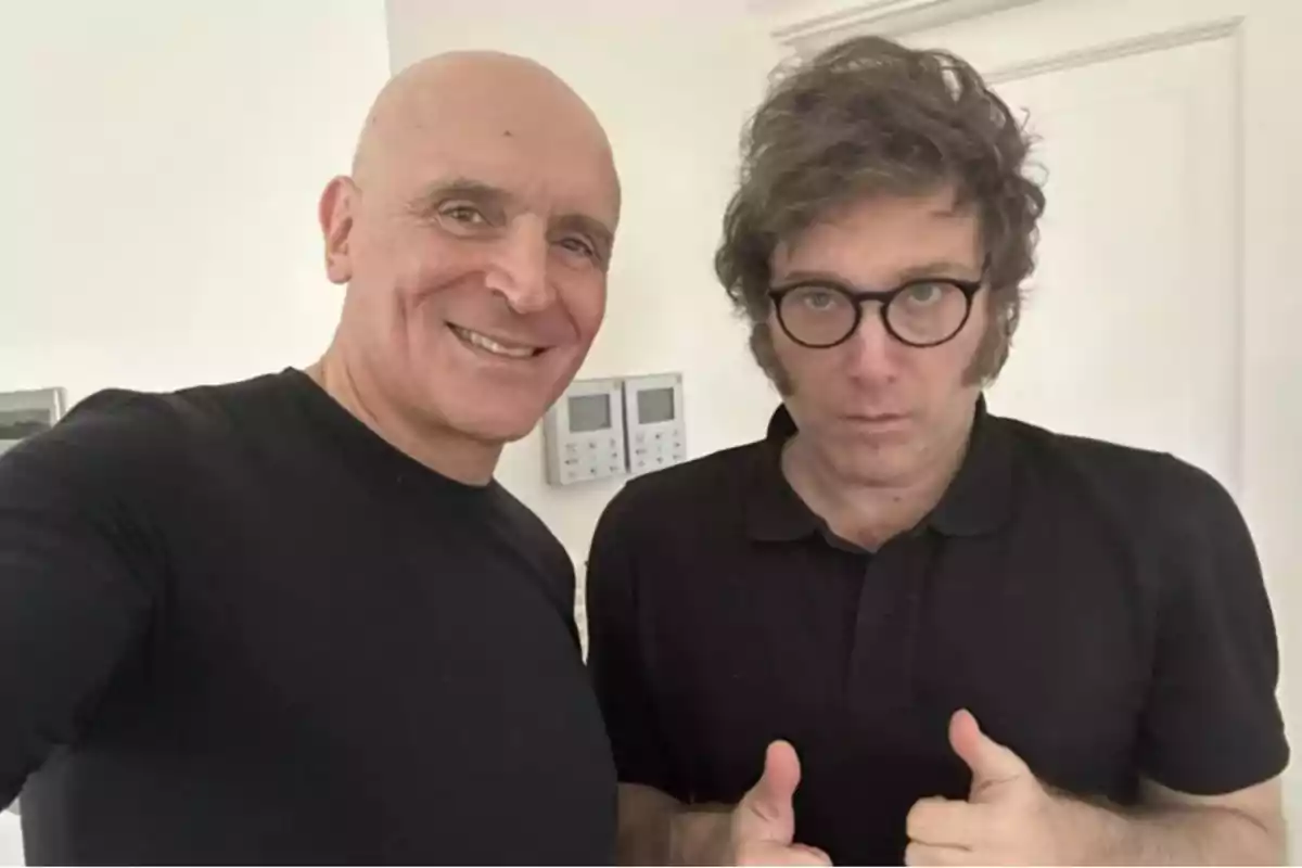 Two men pose together in a well-lit room, one smiling and the other showing a serious expression with both thumbs up.