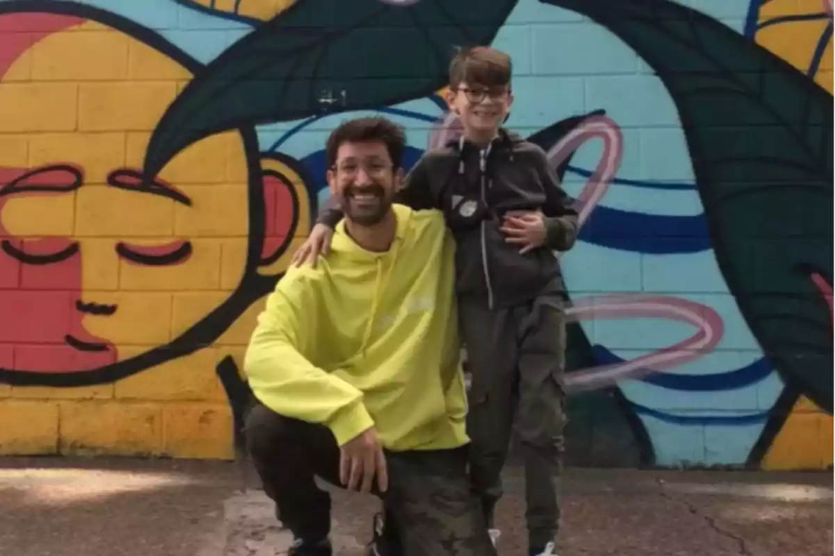 A man and a boy smile in front of a colorful mural with a drawing of a yellow face.