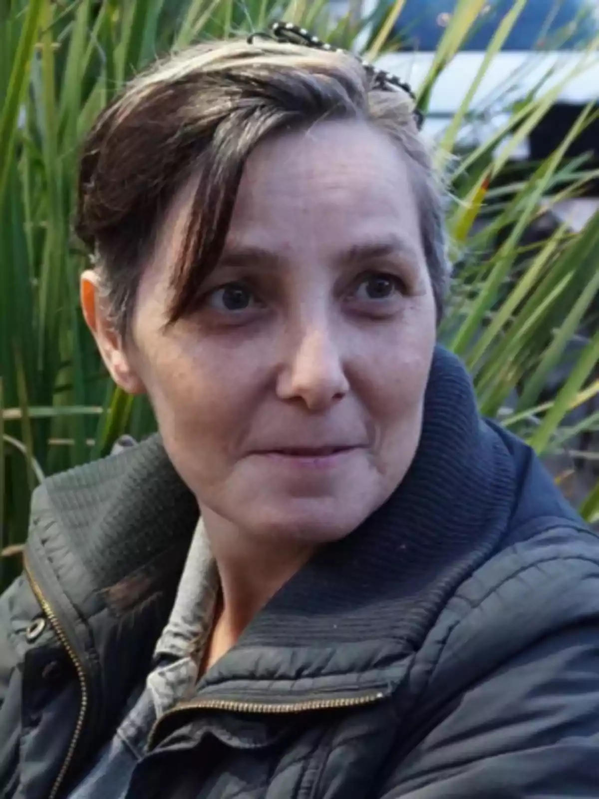 Short-haired, gray-haired woman in a dark jacket sitting outdoors in front of green plants Short-haired, gray-haired woman in a dark jacket sitting outdoors in front of green plants