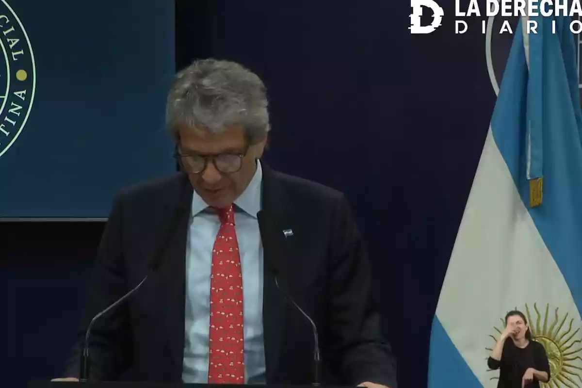Gray-haired man with glasses speaking at a podium with an Argentine flag and a sign language interpreter to the right Gray-haired man with glasses speaking at a podium with an Argentine flag and a sign language interpreter to the right